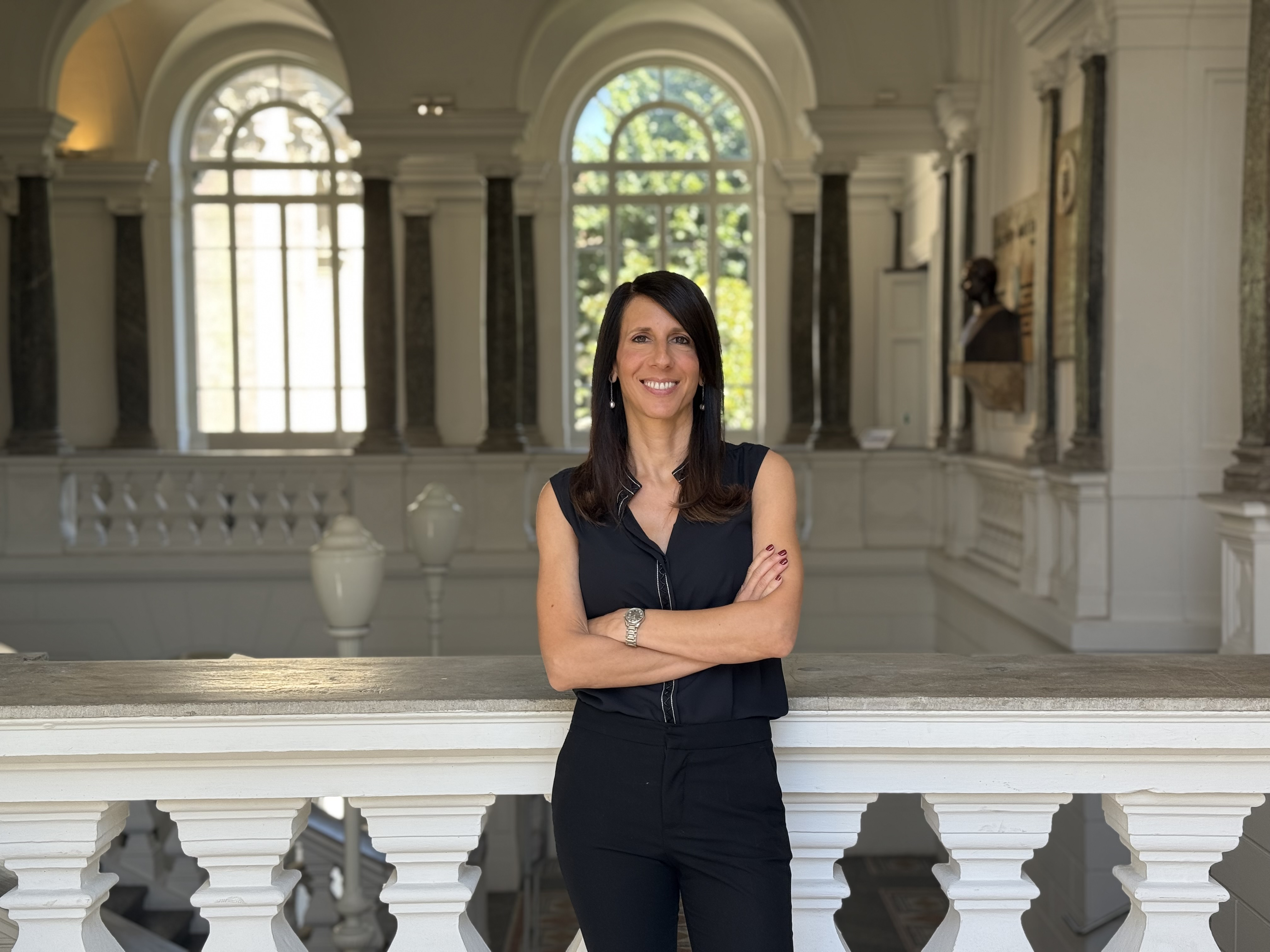 Donna dalla carnagione chiara,mora con capelli di media lunghezza con cchi marroni e sorridente. indossa degli orecchini pendenti, un orologio e una camicia senza maniche nera. sullo sfondo, delle finestre ad arco, una balconata interna sui toni del bianco. 