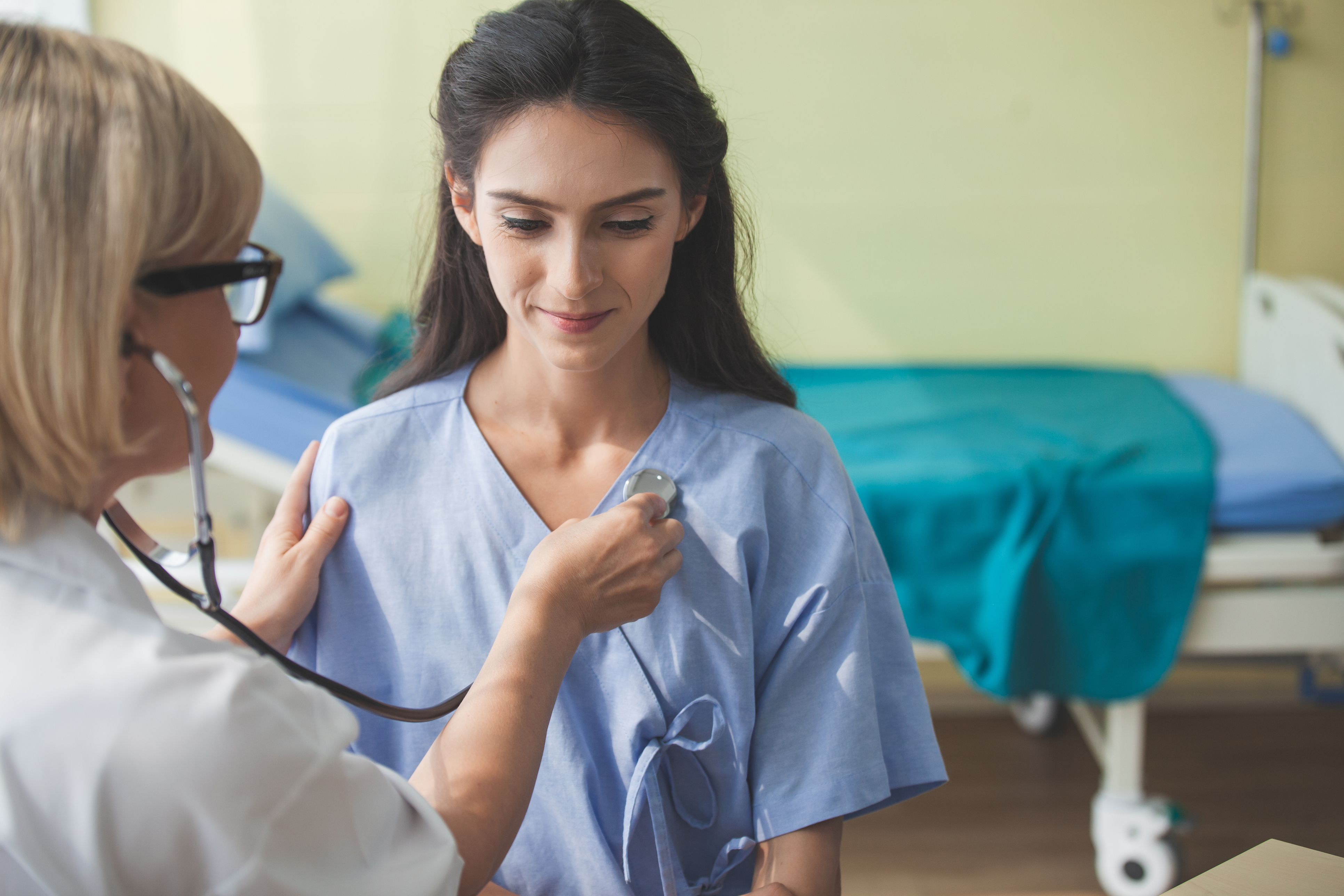 medico di spalle biondo usa lo stetoscopio per sentire il battito di una ragazza magra, con i capelli marroni e una maglietta azzurra. Sullo sfondo una stanza di ospedale e una lettino con un lenzuolo verde 
